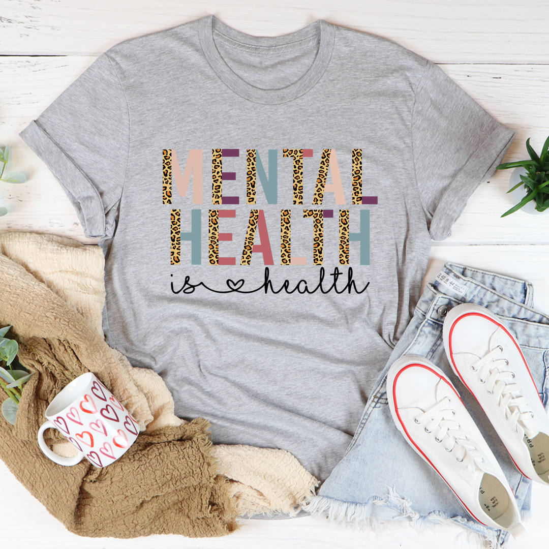 Gray t-shirt with 'Mental Health is Health' text on a white surface with casual items around.