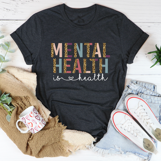 Black t-shirt with 'Mental Health is Health' text on a white surface with casual items around.