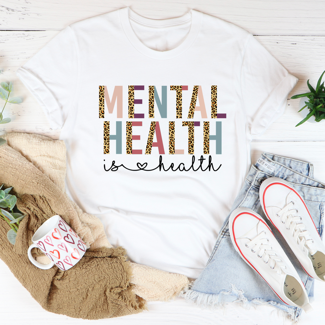 White t-shirt with 'Mental Health is Health' text, mug, scarf, and sneakers on a light wooden surface.