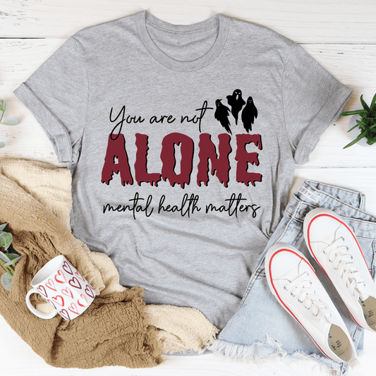 Gray t-shirt with 'You are not alone, mental health matters' text on a white surface with casual items.