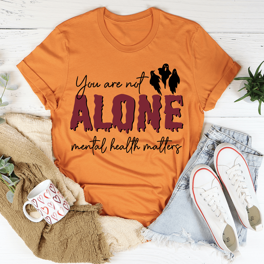 Orange t-shirt with 'You are not alone, mental health matters' text on a white surface with casual items.